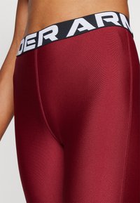 Crvene atletske tajice s crnim elastičnim pojasom na kojem se nalazi bijeli natpis "UNDER ARMOUR". Glatka, prilagođena tkanina s suptilnim sjajem.