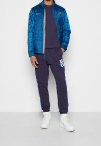 Sininen kuviollinen takki vetoketjulla, laivastonsininen collegepaita, laivastonsiniset collegehousut valkoisilla ja sinisillä yksityiskohdilla sekä valkoiset korkeavartiset tennarit.