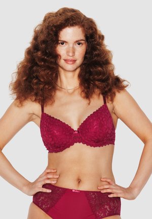 Femme aux cheveux roux bouclés portant un soutien-gorge en dentelle bordeaux assorti et une culotte taille haute, posant les mains sur les hanches devant un fond uni.