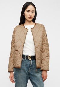 Μπεζ μπουφάν με καπιτονέ ύφασμα, στρογγυλή λαιμόκοψη και καφέ περιγραμματική ρίγα, μπροστινό φερμουάρ, και πλαϊνές τσέπες, σε συνδυασμό με λευκό τοπ και μπλε τζιν.