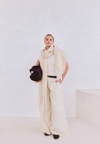 Beige oversized sjaal gedrapeerd over een wit shirt, gecombineerd met high-waisted, crèmekleurige, wijde broekspijpen. Houdt een donkerbruine suède tas vast.