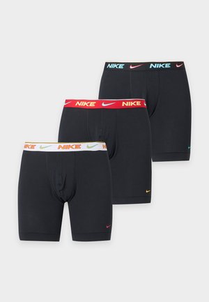 Høj, bar overkrop mand står barfodet iført sorte Nike boxershorts med rød linning mod en ensartet hvid baggrund.