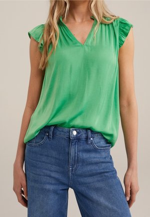 Vrouw die een felgroene mouwloze blouse met gerimpelde schouders draagt, gestopt in een high-waist blauwe jeans, tegen een beige achtergrond.