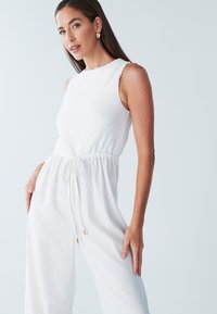 Witte mouwloze jumpsuit met een gestructureerde lijfje, een trekkoord in de taille en wijde pijpen. Bevat gouden accenten op de uiteinden van het trekkoord.