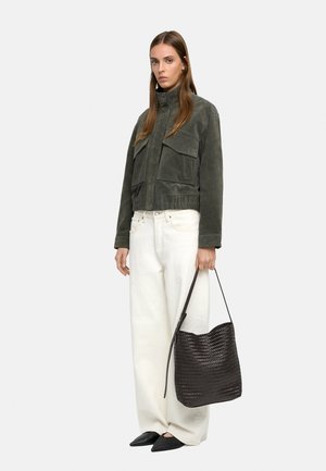 Kobieta ubrana w ciemnozieloną zamszową kurtkę, białe spodnie o szerokich nogawkach, czarne spiczaste buty, trzymająca dużą czarną plecioną torbę na ramię.