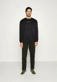 Man standing facing forward wearing a black sweatshirt with text, black pants, and olive green sneakers against a plain white background.