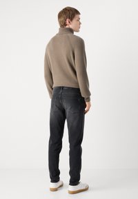 Brown, textured turtleneck sweater with ribbed cuffs, paired with dark jeans and white sneakers. View from the back.