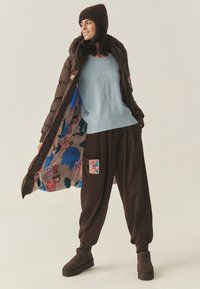 Puchowa brązowa kurtka z kolorową wzorzystą podszewką, jasnoniebieski sweter z motywem misia, brązowe luźne spodnie i brązowe botki za kostkę.