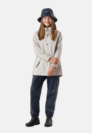Pige iført lysegrå regnjakke, marineblå vandtætte bukser, sorte gummistøvler og marineblå bøllehat, stående med armene krydsede.
