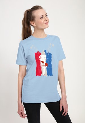 Jonge vrouw met paardenstaart draagt een lichtblauw T-shirt met vintage cartoonfiguur Betty Boop over verticale strepen in rood, wit en blauw.