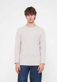 Ανοιχτό μπεζ πουκάμισο μακρυμάνικο με ριμπ υφή και στρογγυλή λαιμόκοψη, με διακριτικό λογότυπο "BOSS" στο στήθος. Φορεμένο με σκούρο μπλε τζιν.