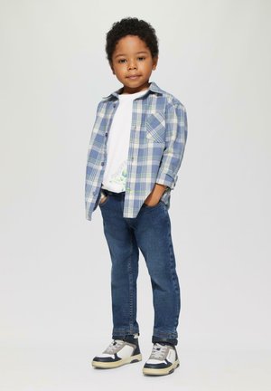 Blauwe en groene geruite overhemd over een wit T-shirt, gedragen met donkere spijkerbroek en multifunctionele sneakers.