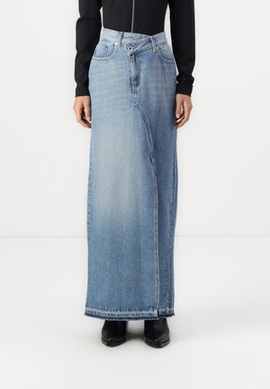 Model draagt een lange, lichtblauwe spijkerrok met asymmetrische voornaad en knoopsluiting, gecombineerd met zwarte laarzen en een zwarte top.