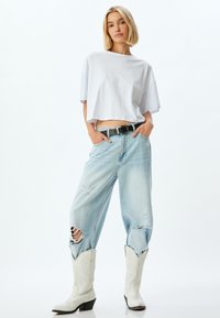 Weißes, kurzes T-Shirt, hellblaue, ausgebleichte Jeans mit Rissen und weiße Cowboy-Stiefel mit spitz zulaufender Zehenpartie und dekorativen Nähten. Schwarzer Gürtel sichtbar.