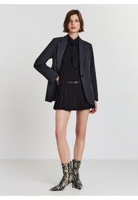 Blazer noir ajusté sur un chemisier noir avec un nœud, associé à une jupe plissée noire et des bottines à imprimé serpent. Accessoires simples.