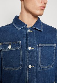 Giubbotto in denim blu scuro con colletto classico, due tasche frontali, bottoni argentati e una texture leggermente vissuta con dettagli di cucitura visibili.