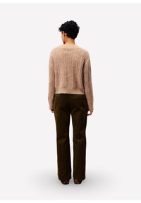 Persoon met kort, krullend haar draagt een beige geribde trui, donkerbruine corduroy broek en schoenen met een luipaardprint, staand met de rug naar ons toe.