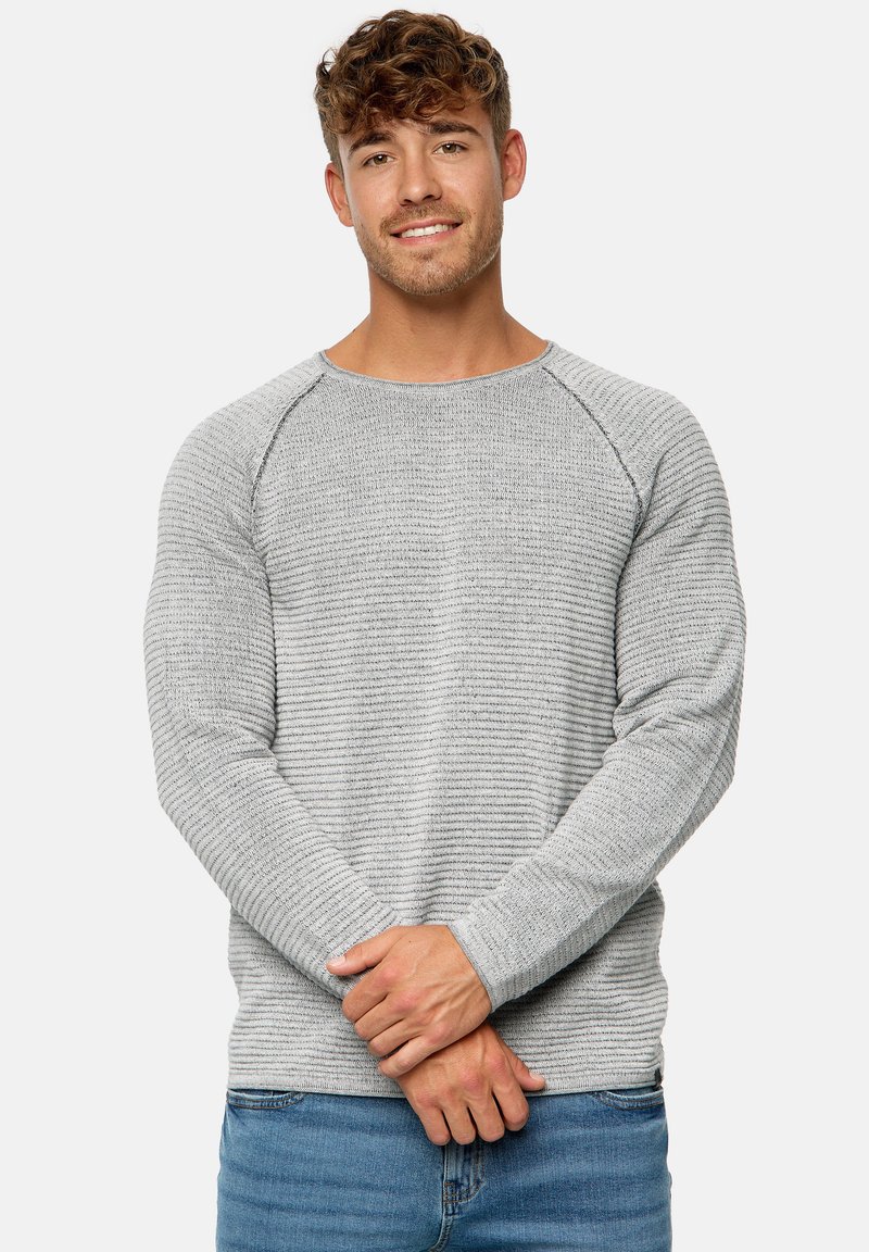Lys grå tekstureret sweater med vandrette striber, rund hals og raglanærmer, parret med blå jeans. Hænderne er samlet foran.