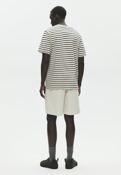 Homme debout de dos portant une chemise à manches courtes rayée blanche et noire, un short beige, des chaussettes grises et des baskets foncées.