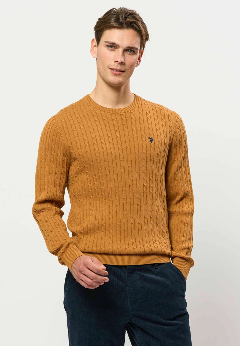 Jeune homme portant un pull torsadé moutarde et un pantalon bleu foncé, debout devant un fond clair uni.