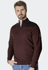 Bordeauxfarbener Reißverschluss-Pullover aus strukturiertem Strick, mit kontrastierenden schwarzen Schulterakzenten, gerippten Bündchen und Saum. Modell trägt ihn mit Denim.