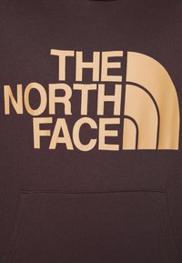 Sudadera marrón con un gran logo beige que dice "THE NORTH FACE" en el pecho; cuenta con un bolsillo frontal y puños acanalados.