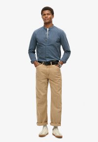 Blauw denim overhemd met lange mouwen en een mandarin kraag, beige opgerolde broeken en witte sneakers met beige accenten. Zwarte riem en polshorloge.