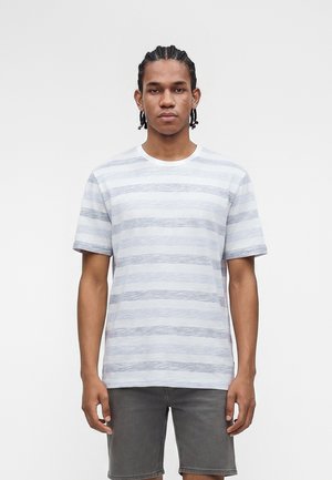 Νέος άνδρας με πλεξούδες, που φοράει λευκό και γκρι ριγέ κοντομάνικο πουκάμισο και σκούρα γκρι σορτς, στέκεται μπροστά σε απλό φόντο.