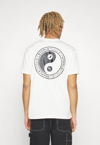 Bílý bavlněný tričko s černým designem yin-yang, na kterém jsou ptáci a text vzadu. Krátké rukávy a kulatý výstřih.
