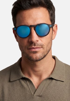 Uomo con capelli corti scuri che indossa occhiali da sole tondi con lenti specchiate blu e una camicia marrone con colletto davanti a uno sfondo semplice.