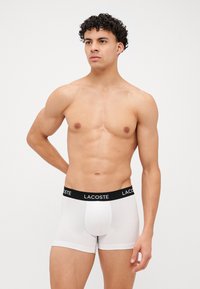 Bele boksarice z črnim pasom, na katerem je logotip 'LACOSTE', narejene iz mehkega, raztegljivega materiala za udobje in prileganje.