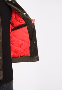 Khaki corduroy jas met een gevoerde rode binnenkant, voorzien van drukknoopsluitingen en een zijzak, gedeeltelijk open weergegeven.