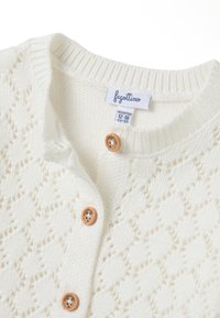 Weißer Strickcardigan mit einem Rautenmuster, ausgestattet mit Holzknöpfen und einem gerippten Kragen. Größenetikett weist auf 12-18 Monate hin.