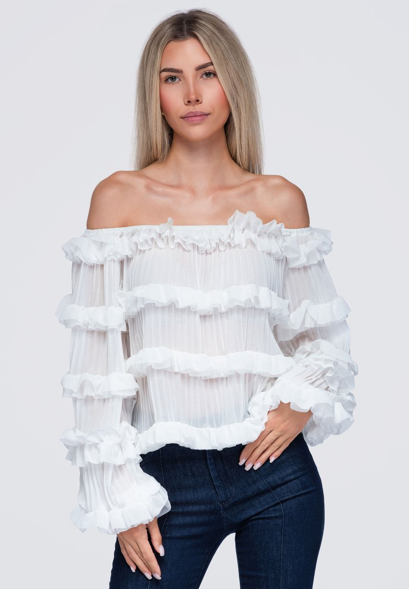 Femme aux longs cheveux blonds portant un chemisier blanc à volants et épaules dénudées ainsi qu'un jean foncé, posant avec une main sur la hanche devant un fond blanc.