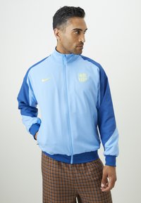 Casaco Nike azul claro e marinho com fecho éclair na frente, punhos canelados e logótipo bordado. O material parece suave e atlético.