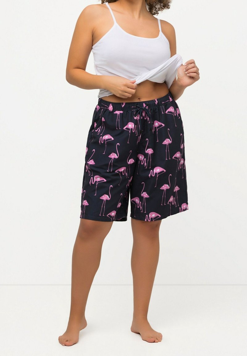 Donna che indossa pantaloncini neri con stampa di fenicotteri rosa e una canotta bianca, alzando leggermente la canotta per mostrare l'elastico dei pantaloncini.