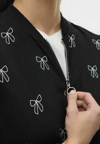 Svart bomberjacka med vita rosett-mönster, försedd med en silverfärgad dragkedja och ribbad krage. Slät tygtextur, närbild av framdelen.