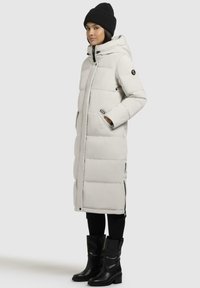 Lang, lysgrå pufferjakke med hette, sidelommer og svarte detaljer, sammen med svarte knelang støvler og en svart strikket lue.