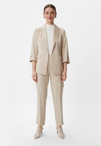 Beżowy garnitur na miarę z jednobutonową marynarką, dopasowanymi spodniami i białym topem z wysokim kołnierzem. Całość dopełniają sandały na obcasie z siateczki.