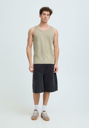 Young man with curly hair wearing a beige tank top, black knee-length shorts, white socks, and beige sneakers standing against a light background.