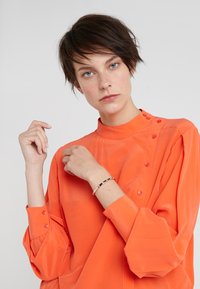 Orange blouse with high collar, button details running along the shoulder, and gathered sleeves. Model wearing a simple metallic bracelet.