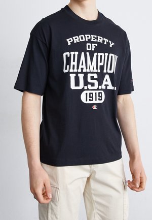 Person iført en sort T-shirt med hvid tekst "PROPERTY OF CHAMPION U.S.A. 1919" og lyse beige cargobukser.