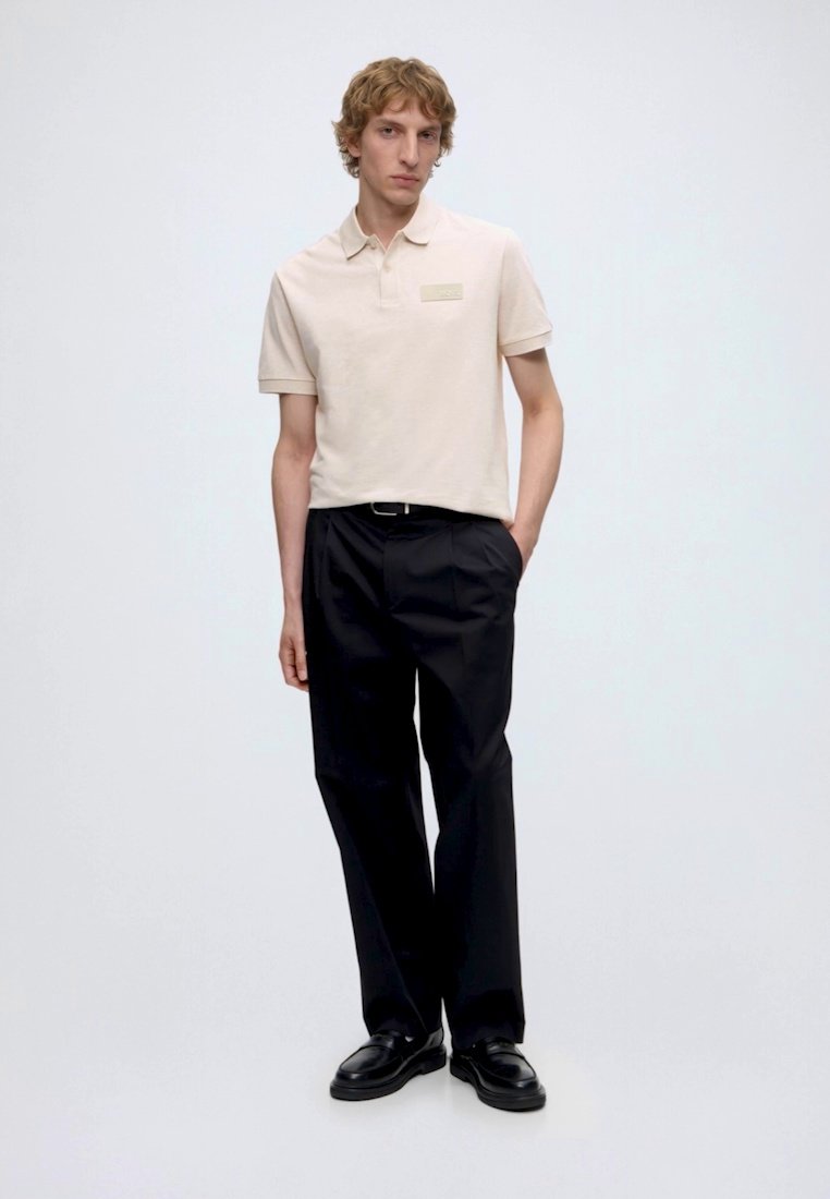 Jeune homme debout, une main dans la poche, portant un polo beige, un pantalon noir et des chaussures noires sur un fond blanc uni.