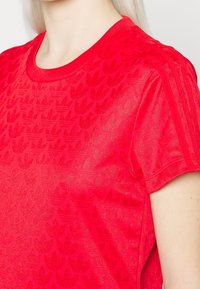 Červené tričko s krátkým rukávem, které má texturovaný květinový vzor. Kulatý výstřih a jemná tkanina zvýrazňují jeho lehký vzhled.