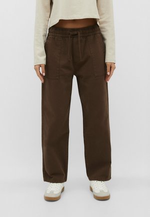 Personne portant un pantalon marron à jambes larges avec des poches frontales, des baskets blanches et un haut court à manches longues beige clair, debout sur un fond uni.