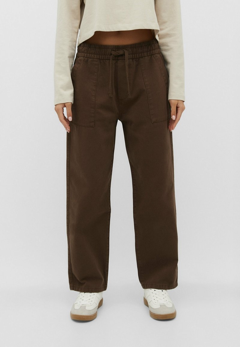 Personne portant un pantalon marron à jambes larges avec des poches frontales, des baskets blanches et un haut court à manches longues beige clair, debout sur un fond uni.