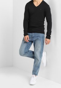 Schwarzes langärmliges V-Ausschnitt-Shirt kombiniert mit hellblauen Jeans und weißen Turnschuhen. Der Stoff wirkt glatt, mit lässigem Schnitt und minimaler Detailgestaltung.