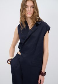 Mörkblå ärmlös kavaj med skarpa slag, sned sidokant och fickdetalj, kombinerad med matchande byxor och två träarmband.