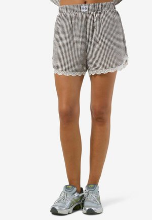 Picioare purtând pantaloni scurți maro și albi cu model în carouri, cu dantelă la margine și pantofi sport Asics argintii, cu accente verde deschis.