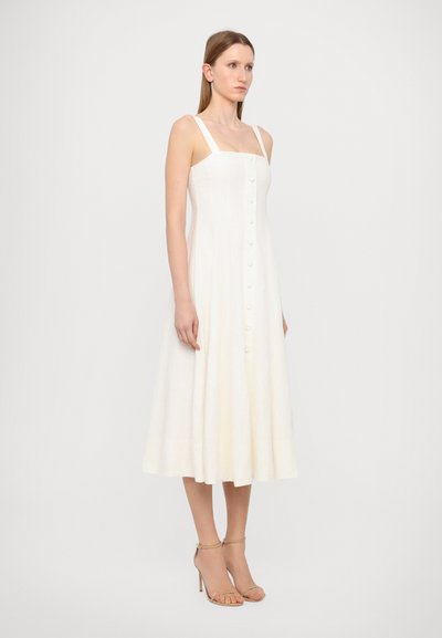 Vestido de lino blanco con escote cuadrado y tirantes anchos, con un frente abotonado y una falda acampanada. Combinado con sandalias de tiras.